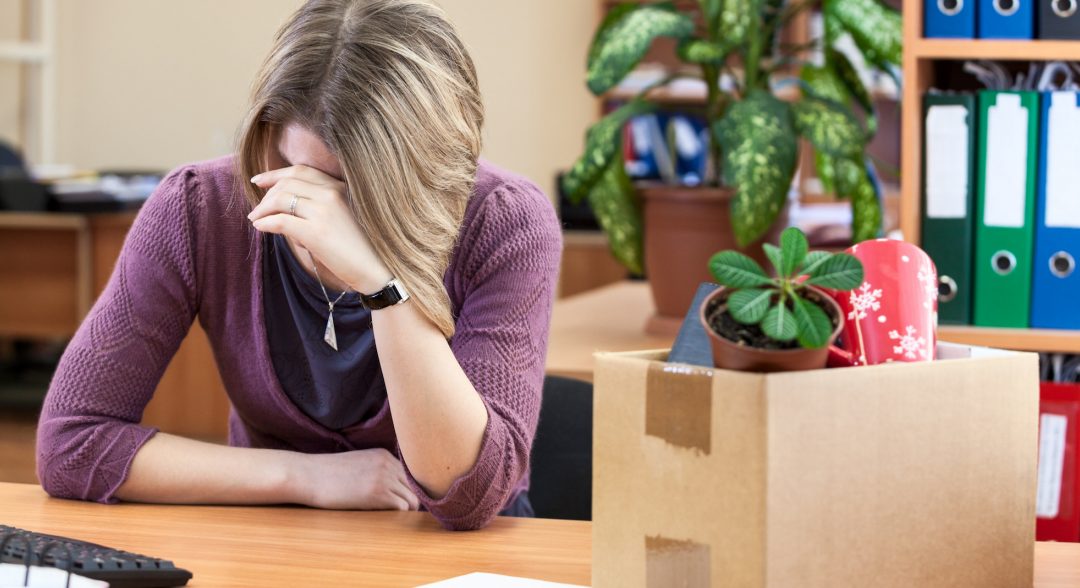 Woman sitting at her desk with her head in her hands. A cardboard box of belongs beside her.